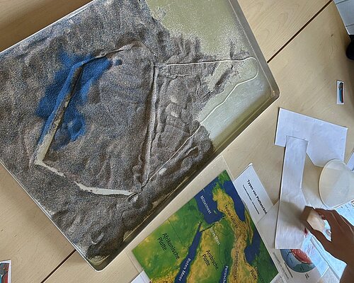 Nachbau der Erdoberfläche mit Sand und Farbe in einem Rahmen.